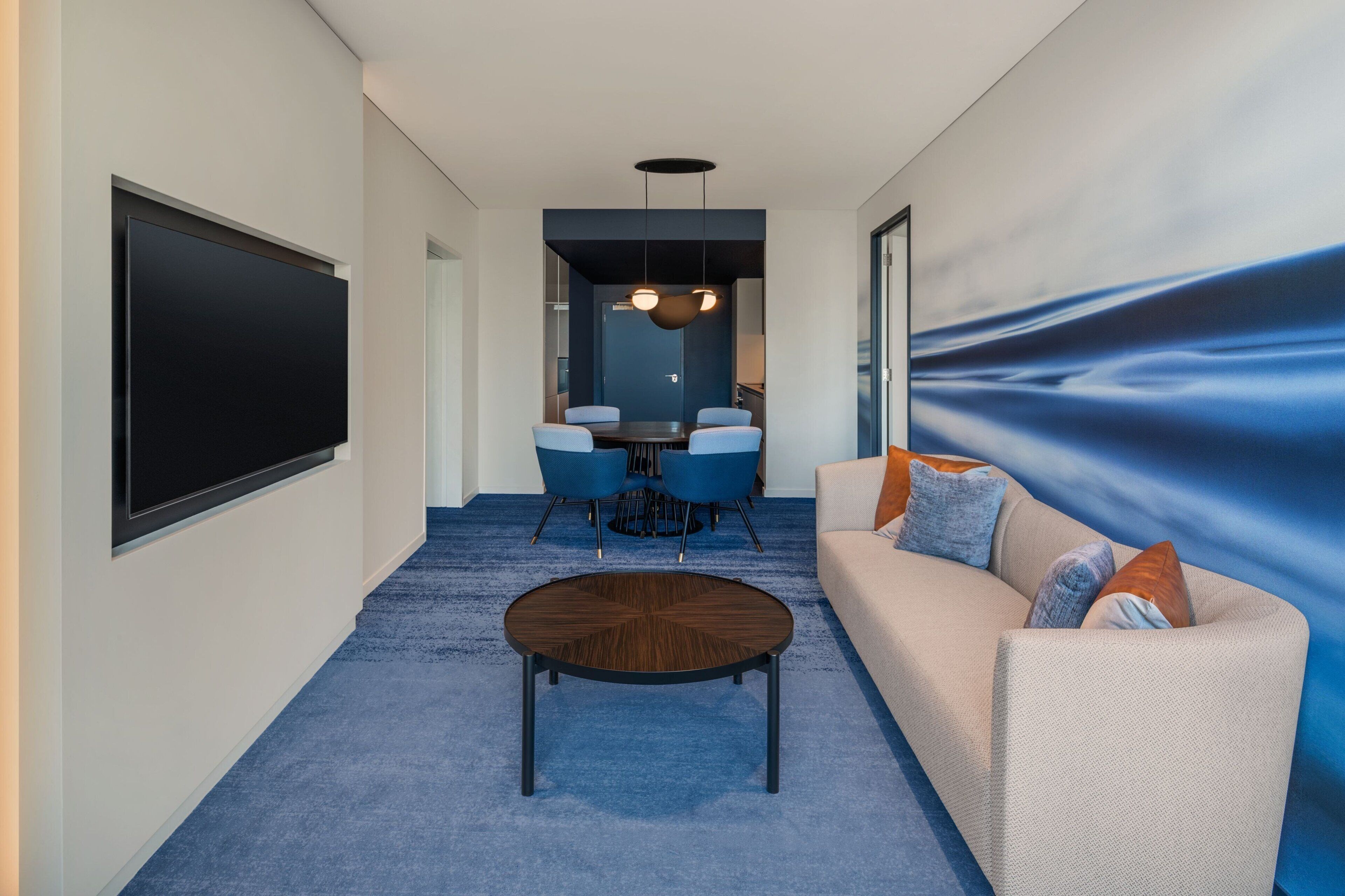 A modern hotel room with a large flat-screen TV, a round coffee table, a sofa with cushions, a dining area with a table and chairs, and a blue carpet.