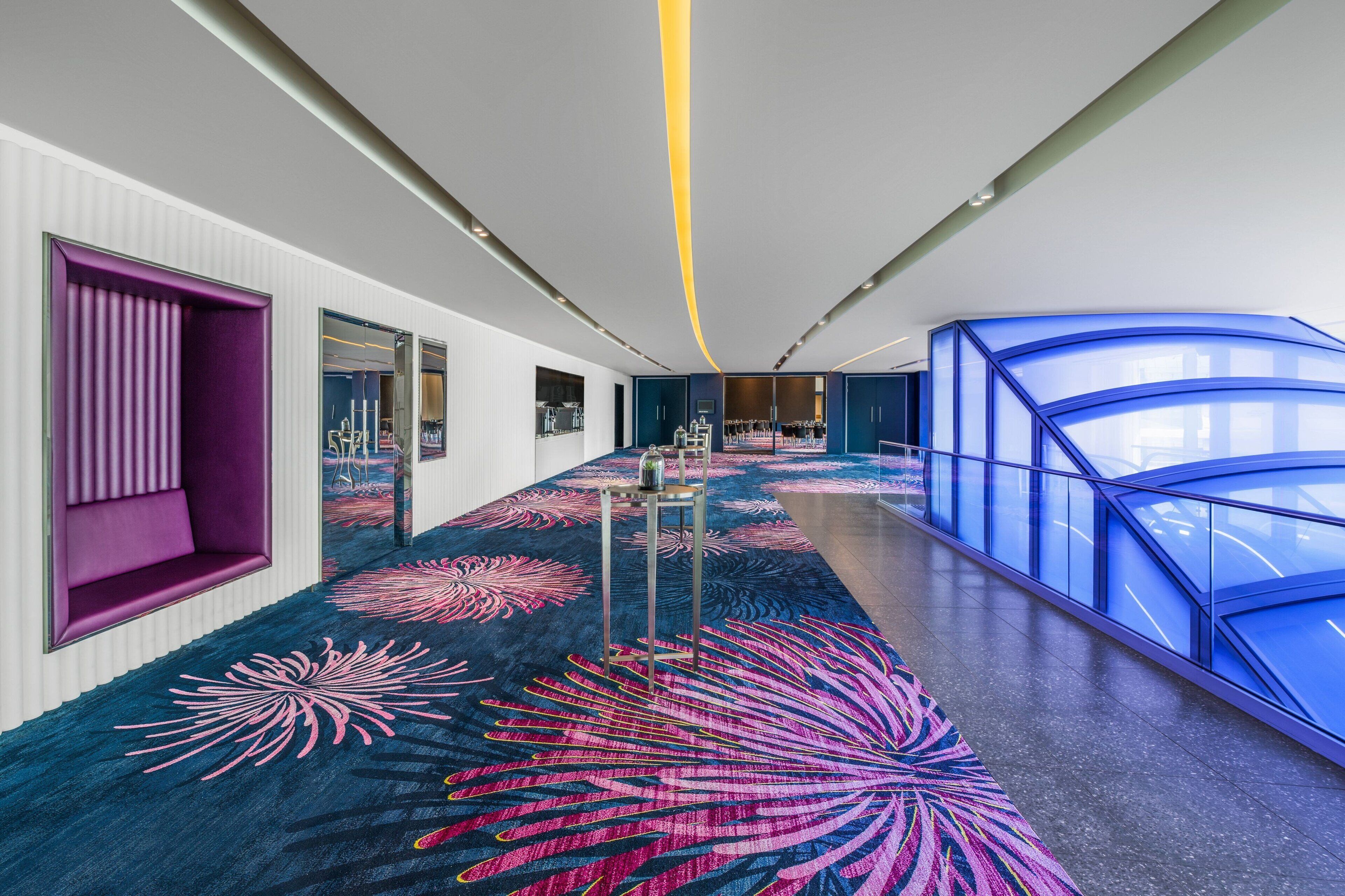A modern hotel corridor with a vibrant carpet, a purple seating area, and a blue glass structure.
