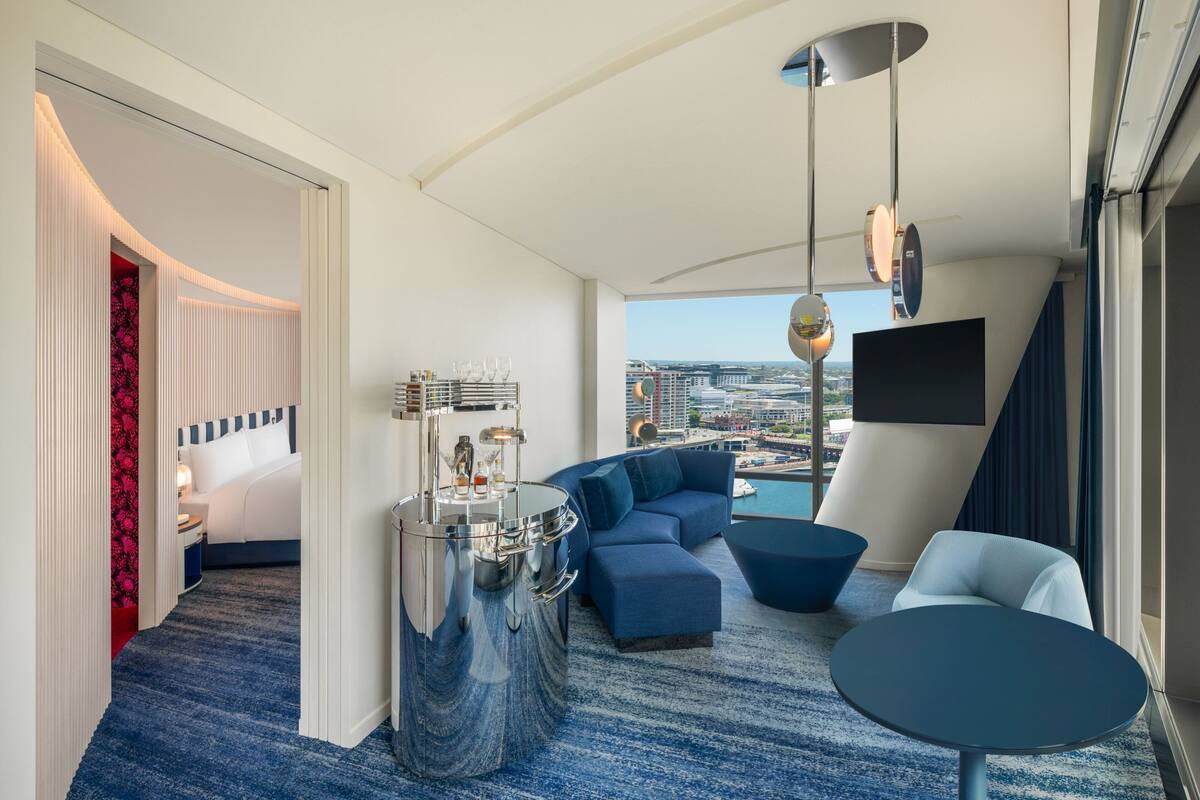 A modern hotel room with a large window, a sofa, a round table, and a minibar.