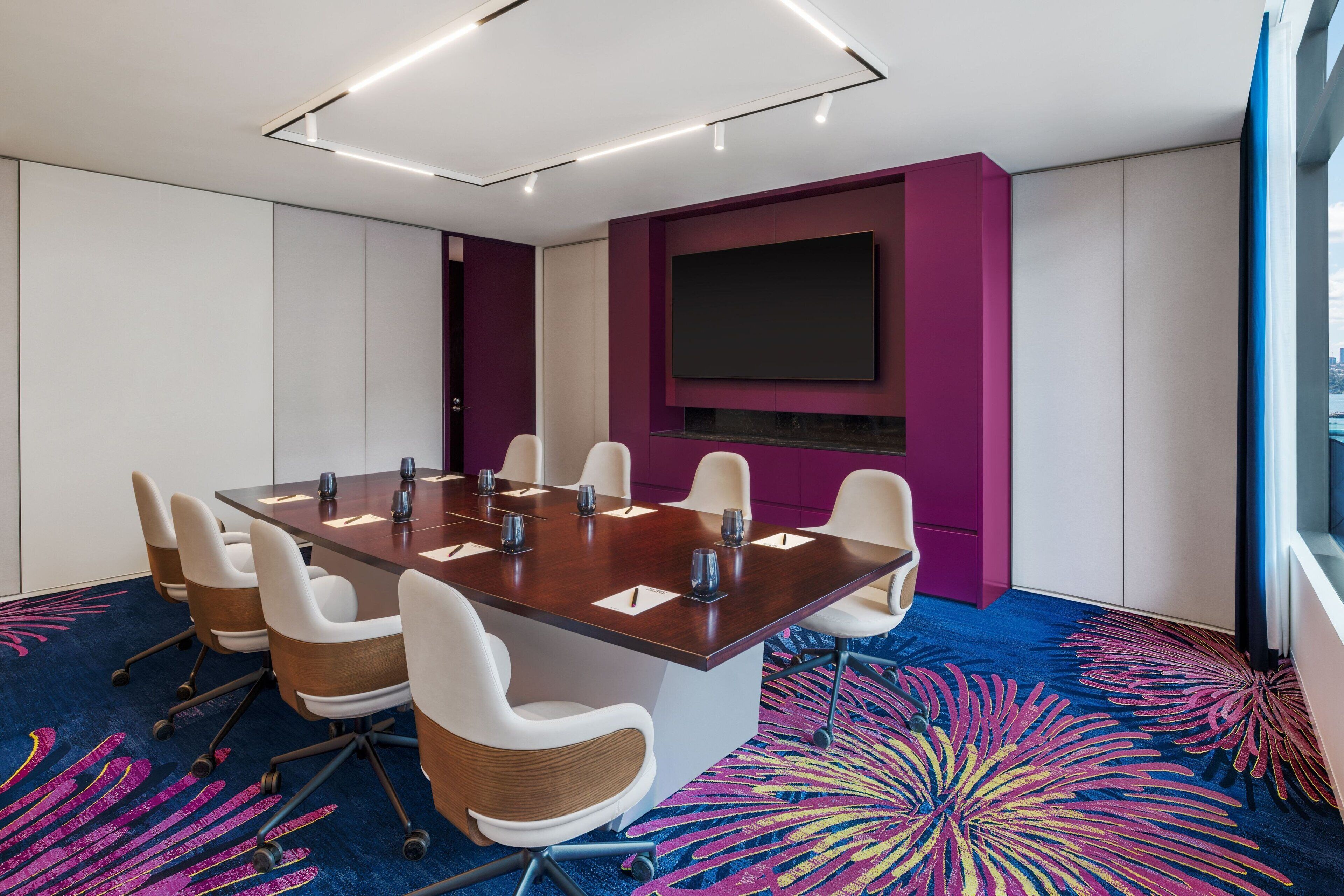 A conference room with a large table, white chairs, a flat-screen TV, and a colorful carpet.