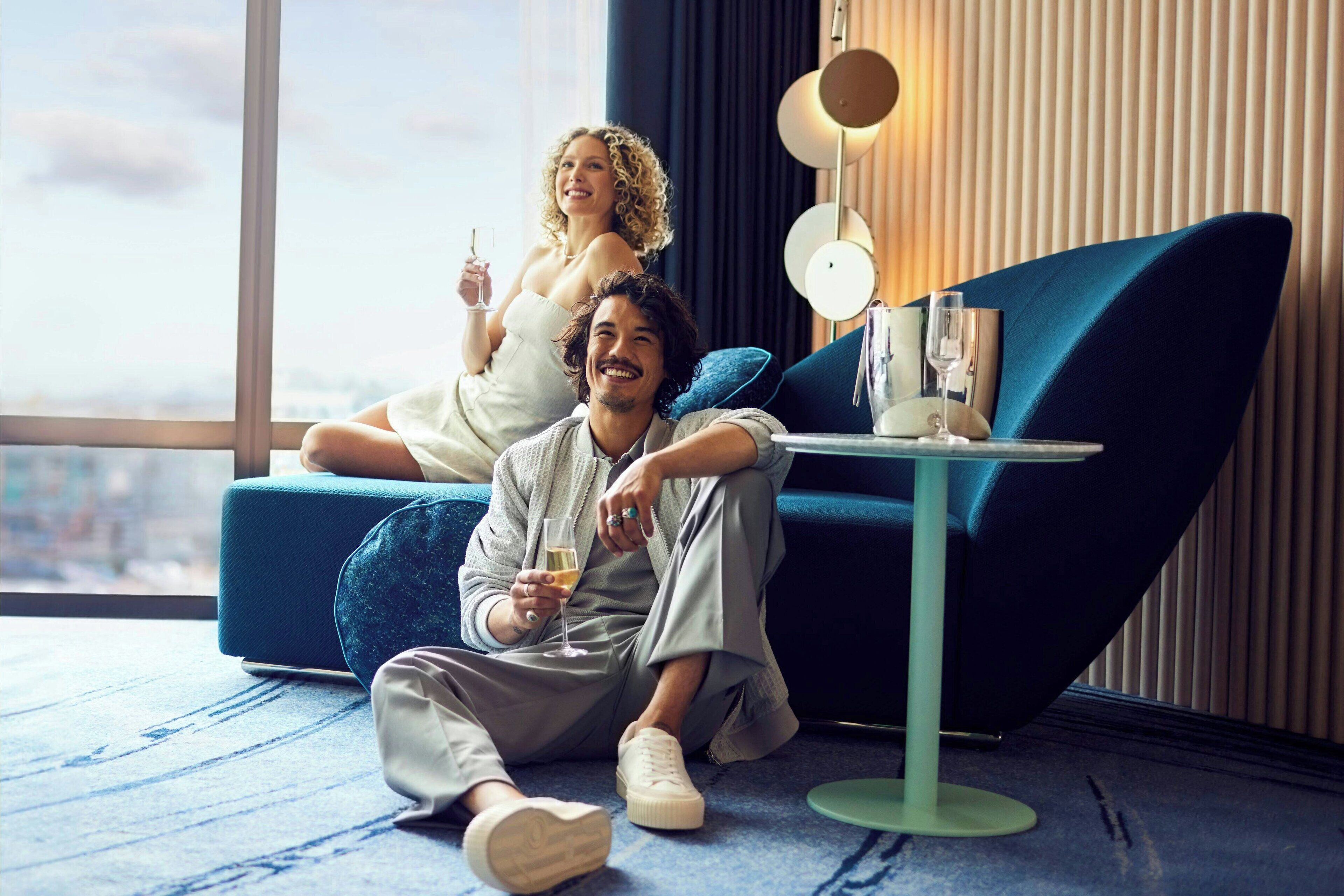 A modern living room with a blue sofa, a small round table with a glass, and a city view through large windows.