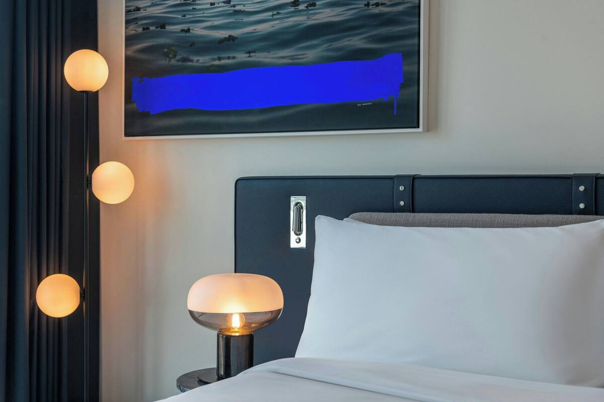 A neatly made bed with white linens, a dark headboard with a metallic handle, and a wall-mounted painting of a blue abstract scene.