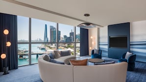 A modern hotel room with a large curved sofa, a small round table, and a flat-screen TV. The room features large windows offering a city skyline view. - W Sydney (Sydney)