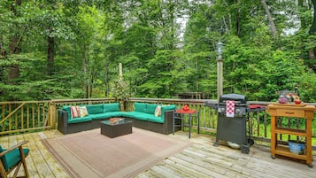 Une terrasse extérieure avec un canapé vert à coussins, un barbecue et une table en bois sur laquelle est posée une bouteille de bière Corona.