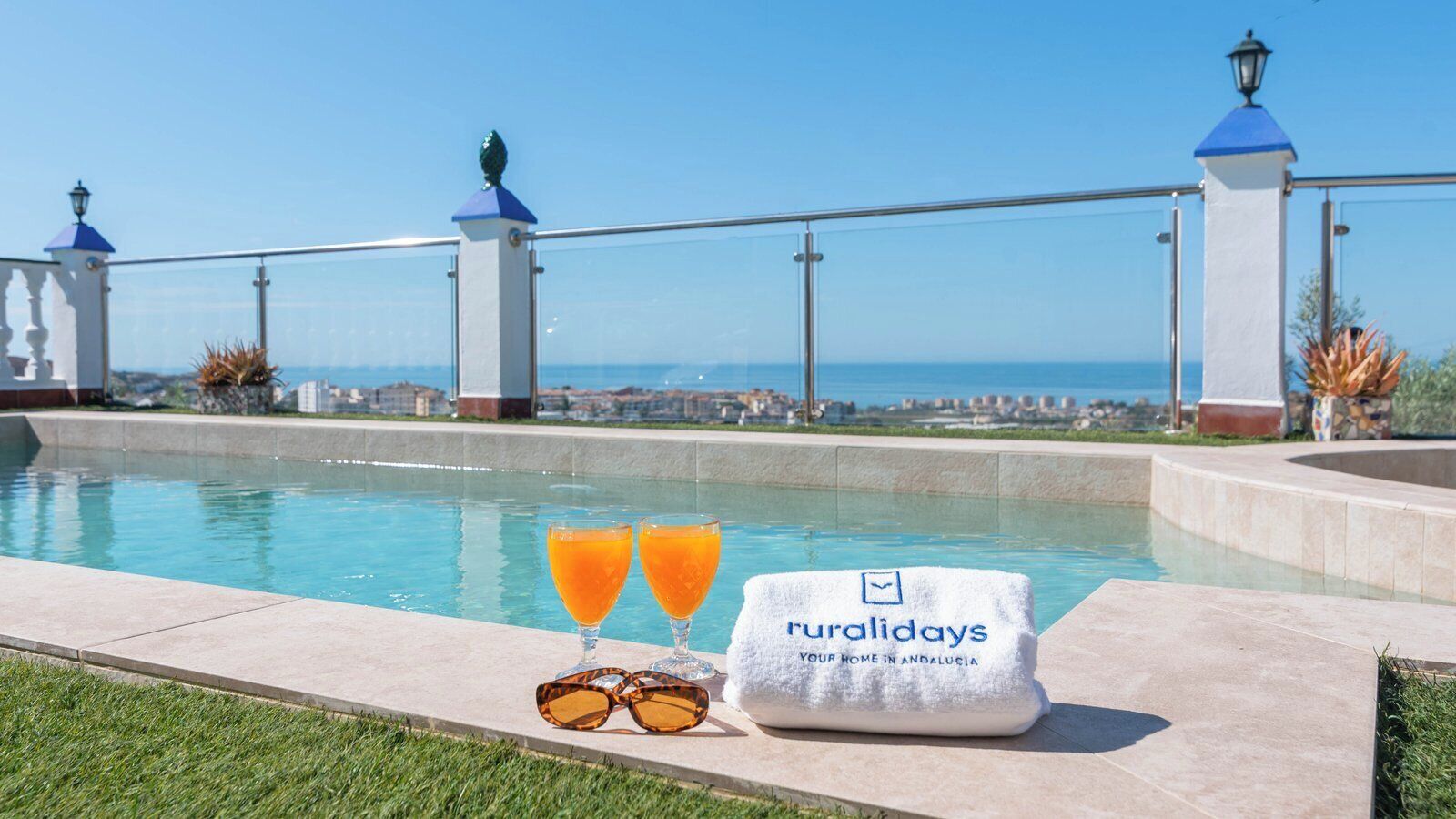 A pool area with a view of the sea, a towel with 'ruralidays' branding, and two glasses of orange juice.