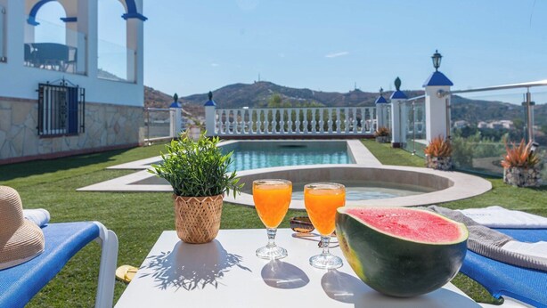 A poolside table with a watermelon, two glasses of orange juice, and a woven basket with greenery. - Casa Mirador Azul Torrox by Ruralidays (Málaga)