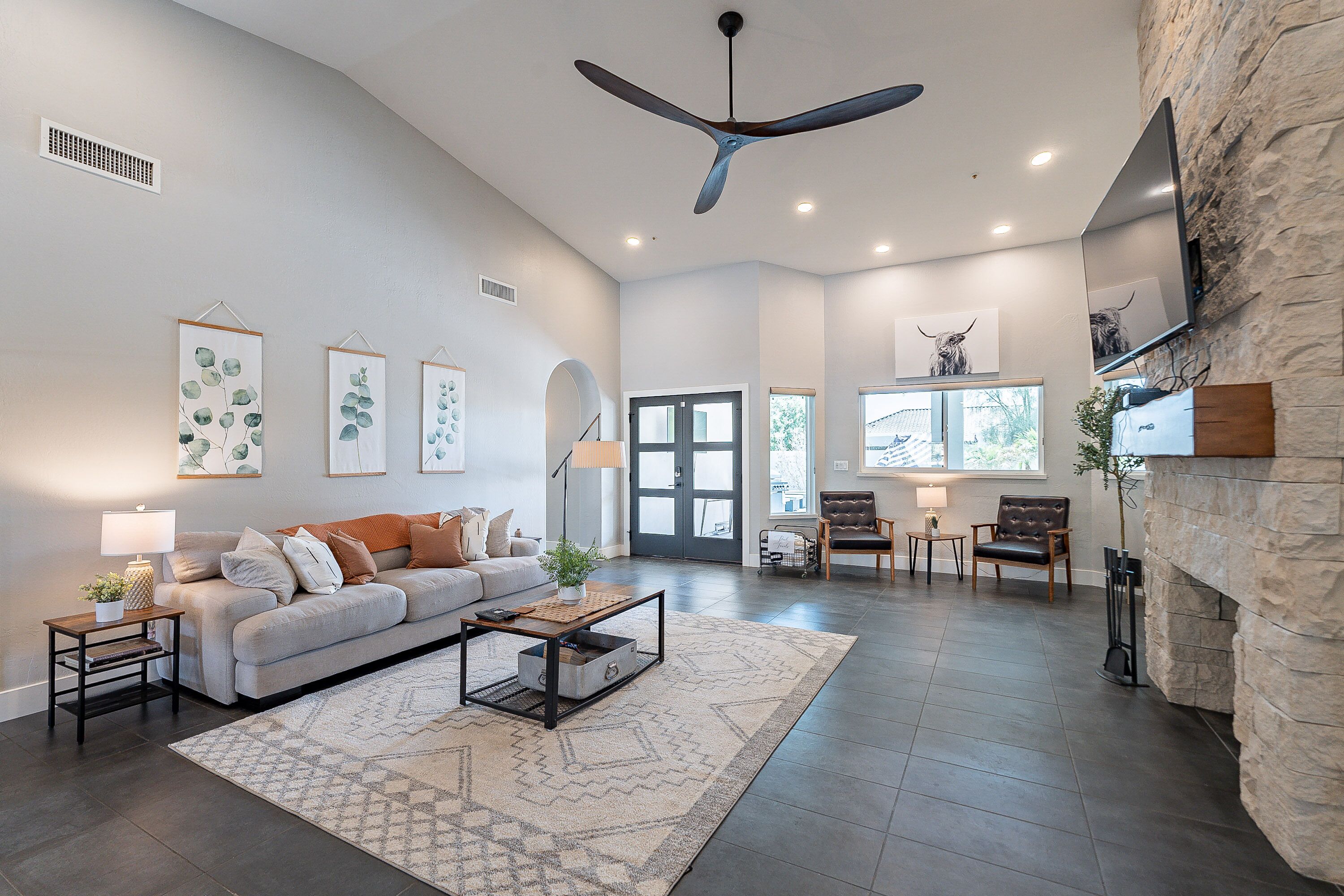 A spacious living room with a high ceiling fan, a fireplace, and a large TV mounted on a stone wall.