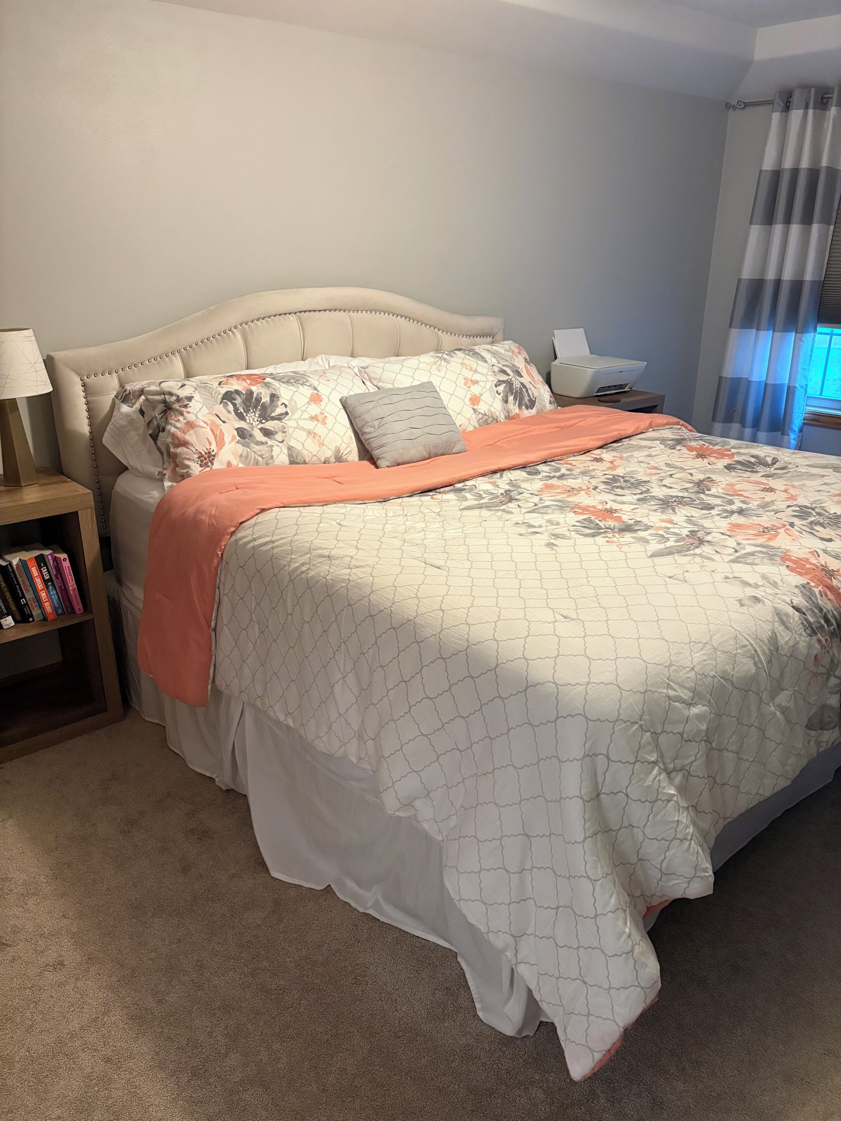 A bedroom with a bed, a nightstand with books, a lamp, and a window with curtains.