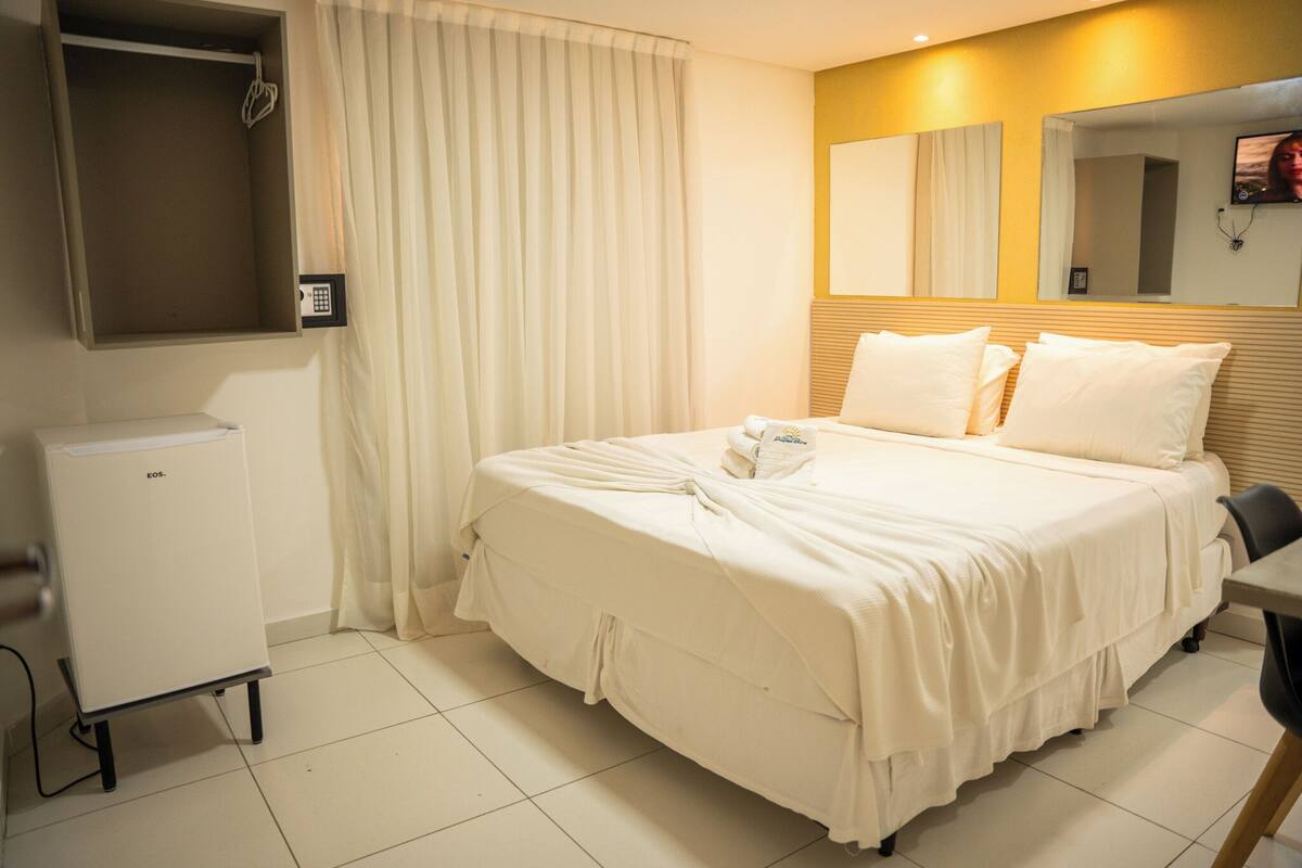 A hotel room with a bed, a small refrigerator, a mirror, and a window with curtains.