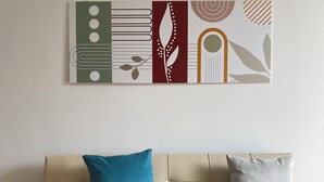 Een beige bank met twee kussens, één turquoise en één grijs, tegen een witte muur met een abstract geometrisch kunstwerk.