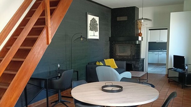 A modern living room with a round wooden table, black chairs, a sofa with yellow cushions, a staircase, and a kitchen in the background. - Maison Proche Centre Ville de Moulins (Yzeure)