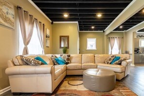 Relax in this stylish living room with a large L-shaped sofa, neutral-toned ottoman, and a kitchenette area.