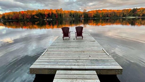 Un molo di legno con due poltrone Adirondack rivolte verso un lago tranquillo, circondato da alberi dai colori autunnali.
