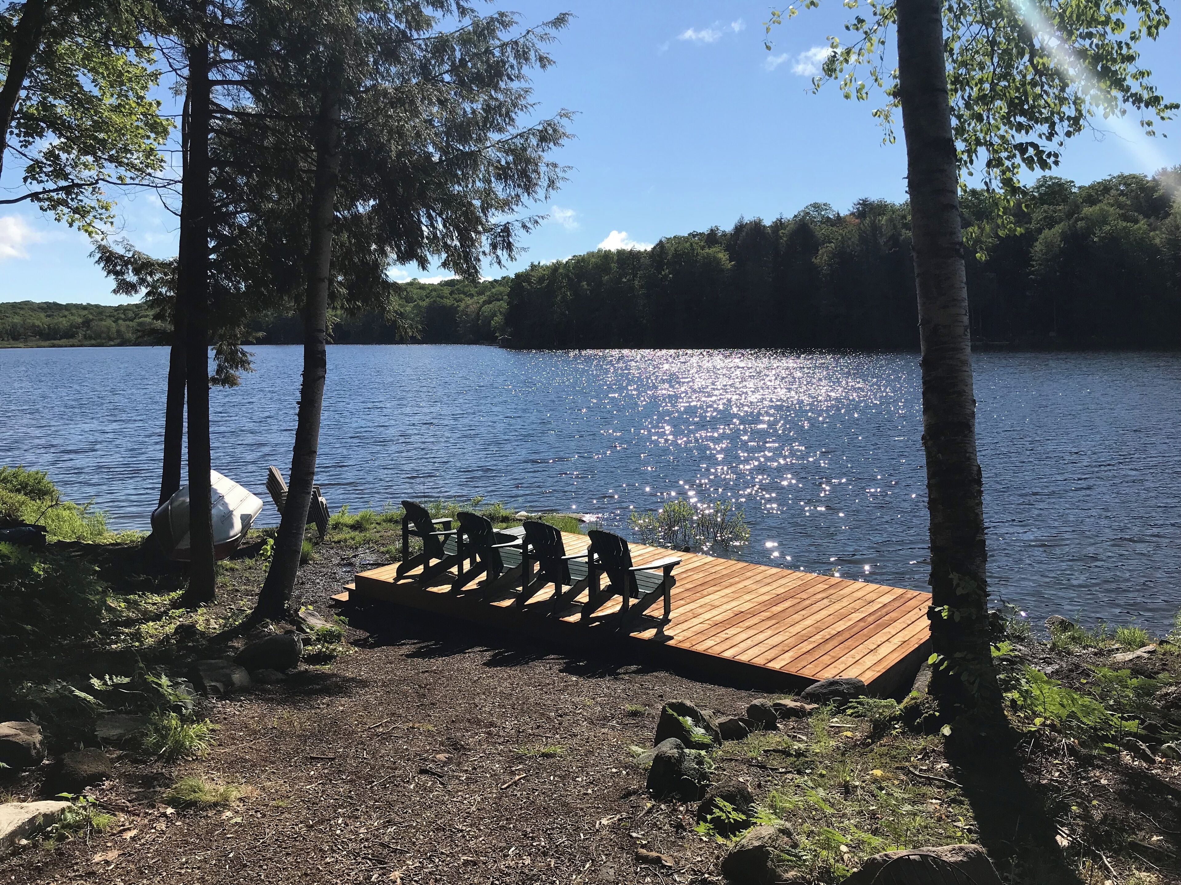 Une terrasse en bois avec des chaises, donnant sur un lac et entourée d’arbres.