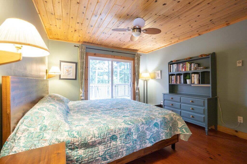 A bedroom with a bed, a ceiling fan, a bookshelf, and a window with a view of trees.