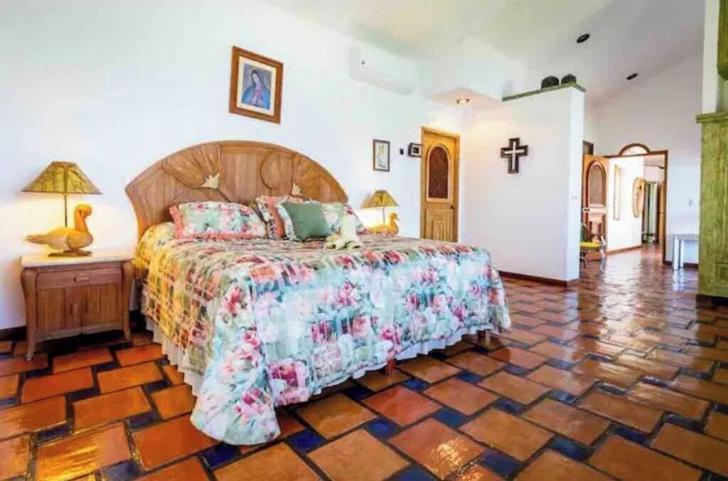 Une chambre à coucher avec un lit en bois, une literie à motifs floraux, une table de chevet avec une lampe et une croix accrochée au mur.