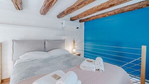 Un dormitorio con cama, almohadas, una mesita de noche con lámpara, una pared con panel azul y barandilla de balcón.