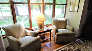 A room with two armchairs, a table with a lamp, and a view of a garden through large windows.