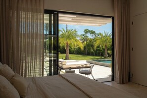 Une chambre avec un grand lit, une porte-fenêtre coulissante donnant sur une terrasse aménagée avec du mobilier d’extérieur et une piscine.