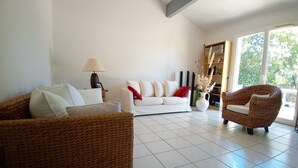 Ein Wohnzimmer mit einem weißen Sofa, einem braunen Korbstuhl und einem weißen Sessel. Auf einem Beistelltisch steht eine Lampe, eine Vase mit getrockneten Pflanzen und ein Bücherregal.