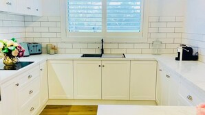 A modern kitchen with white cabinets, a wooden floor, a window with blinds, and a countertop with a coffee machine.