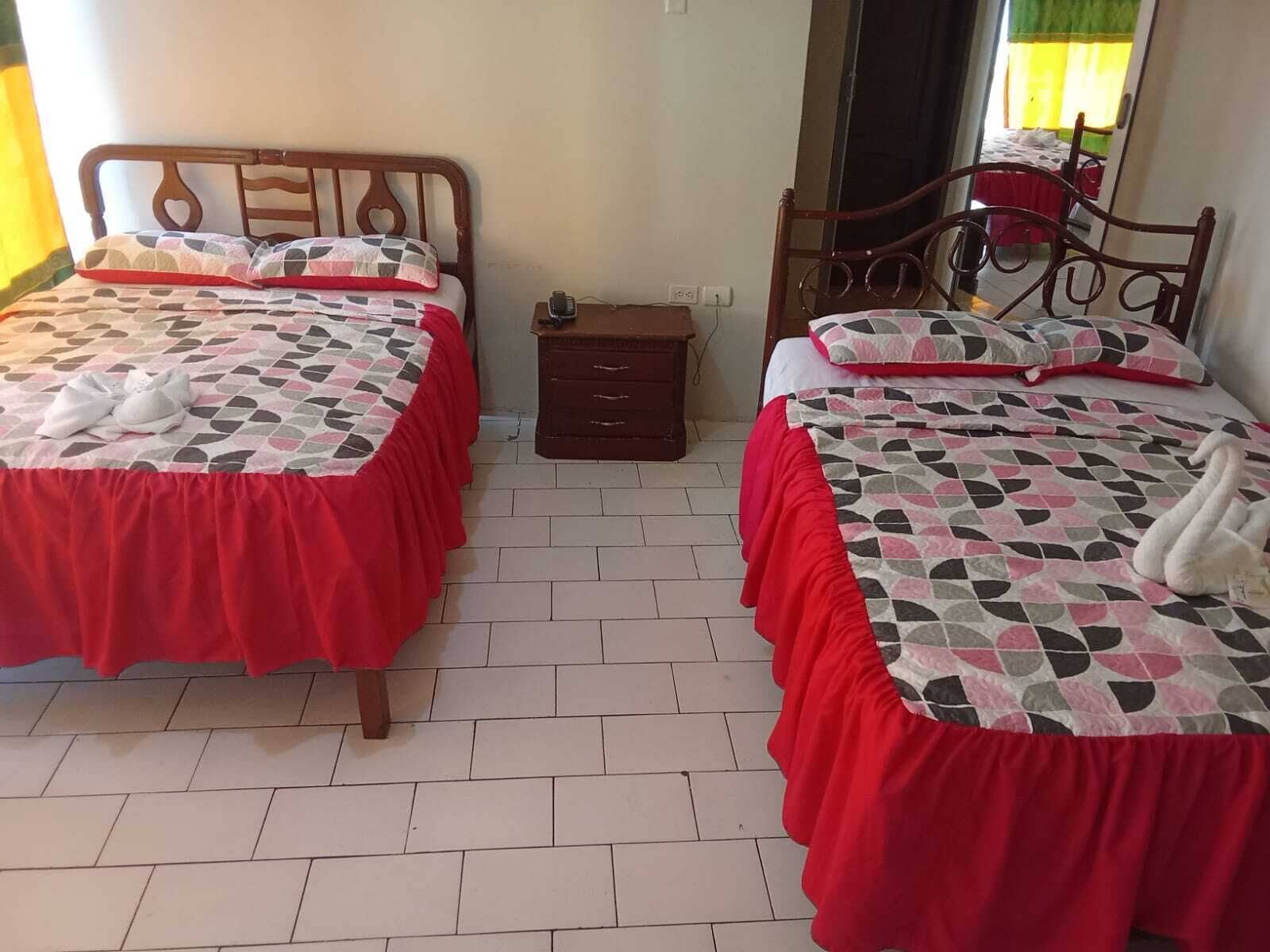 A bedroom with two beds, each with a red bedspread and matching pillows, a wooden headboard, and a small wooden nightstand between the beds.