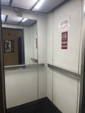 An elevator interior with a mirrored wall, a 'Prohibido Fumar' sign, and a sign indicating a maximum of 5 people.