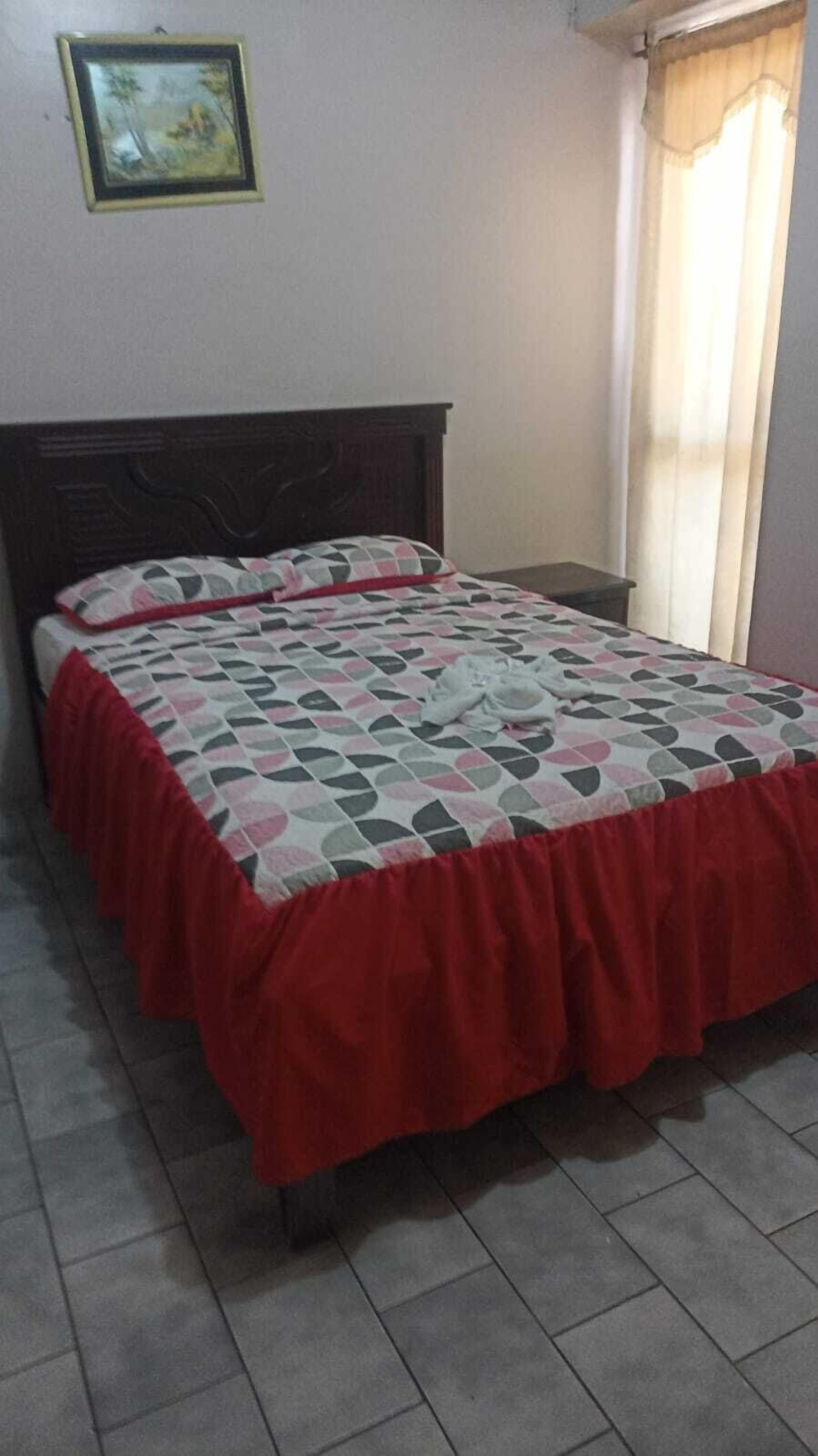 Un lit avec une couvre-lit à motifs et des bordures rouges, une tête de lit en bois et un tableau encadré accroché au mur.