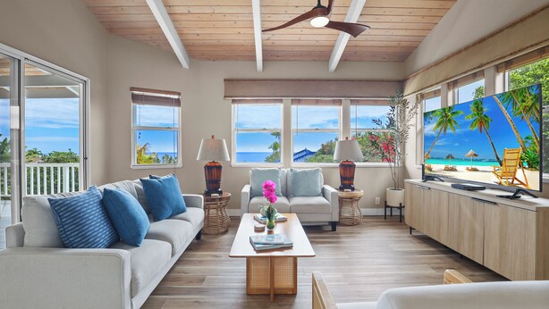 Un salon spacieux avec un plafond en bois, de grandes fenêtres donnant sur la plage, un canapé avec des coussins bleus, une table basse et un téléviseur diffusant une scène de plage.