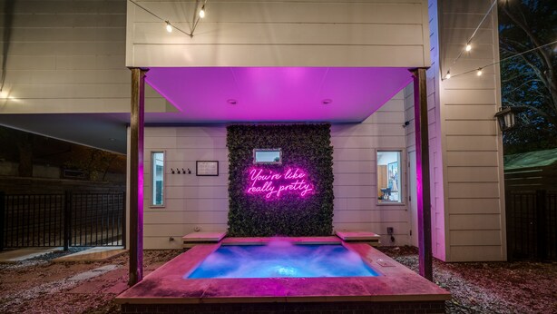Un espace extérieur avec une piscine, agrémenté d’une enseigne au néon, entouré d’une terrasse en bois et d’un mur recouvert de verdure.