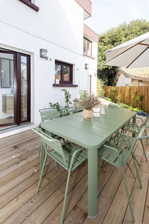 Ein grüner Metalltisch und Stühle auf einer Holzterrasse, im Hintergrund ein weißes Gebäude.