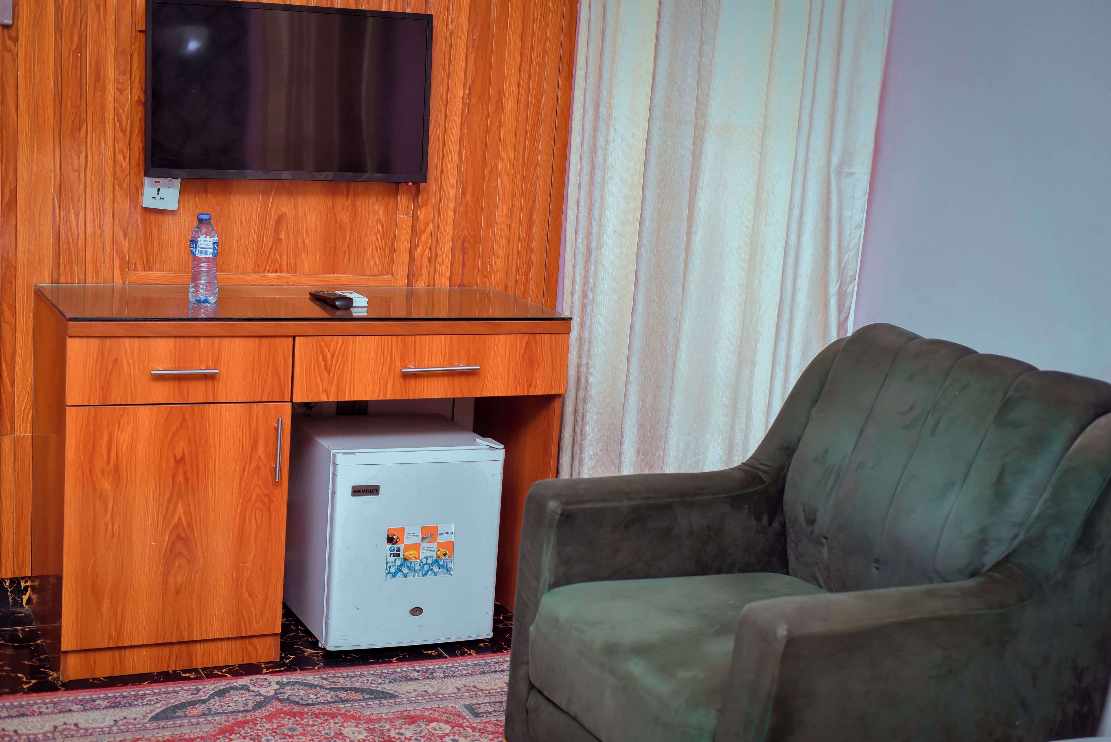 A room with a wooden desk, a small refrigerator, a television mounted on the wall, and a green sofa.