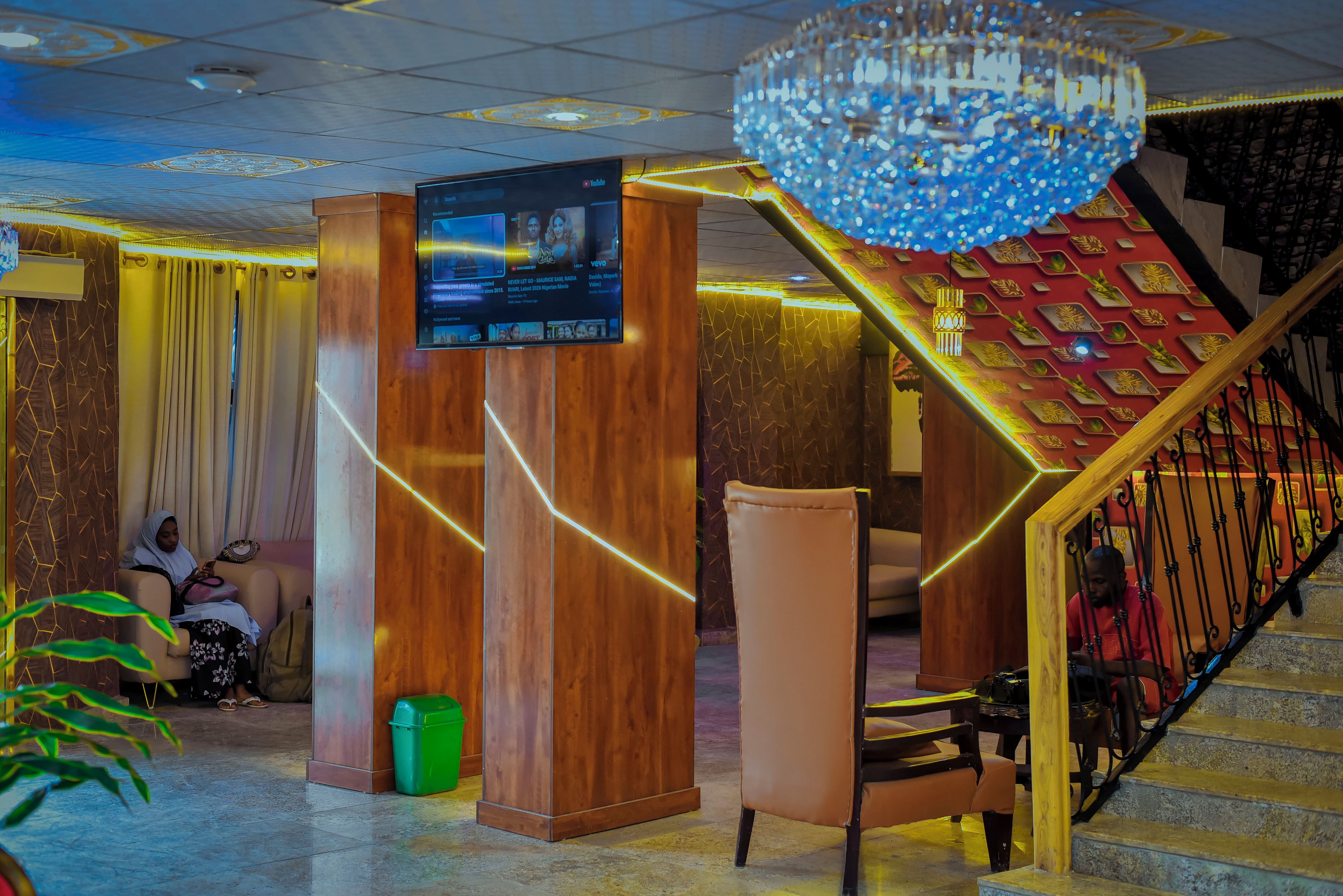 A lobby with a wooden reception desk, a television screen, a staircase with decorative railings, and a chandelier.