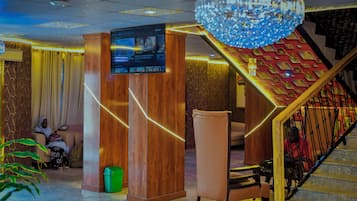 A lobby with a wooden reception desk, a television screen, a staircase with decorative railings, and a chandelier.