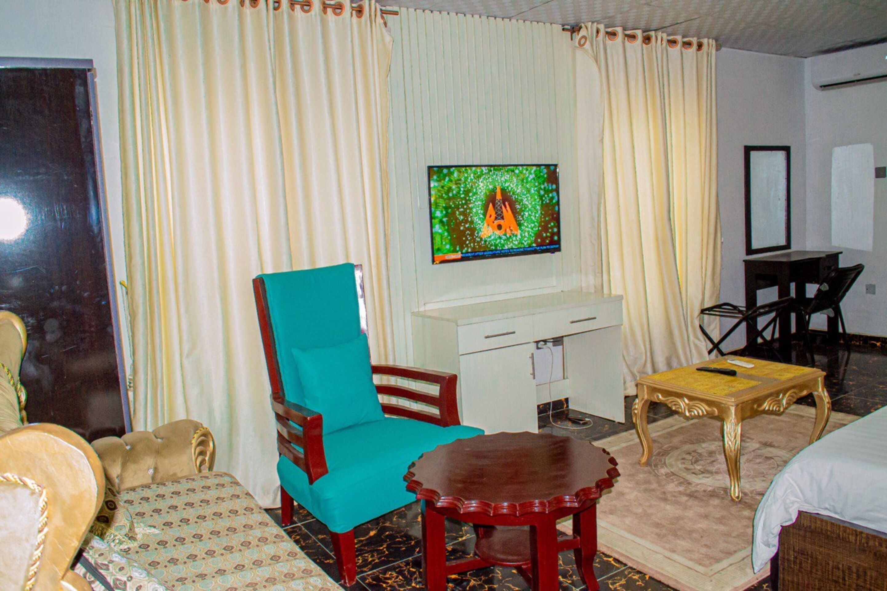 A hotel room with a teal armchair, a wooden table, a TV on the wall, and a bed.