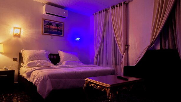 A hotel room with a bed, a nightstand, a lamp, an air conditioner, a painting, and curtains. - Zaytun Hotel Kano (Kano)
