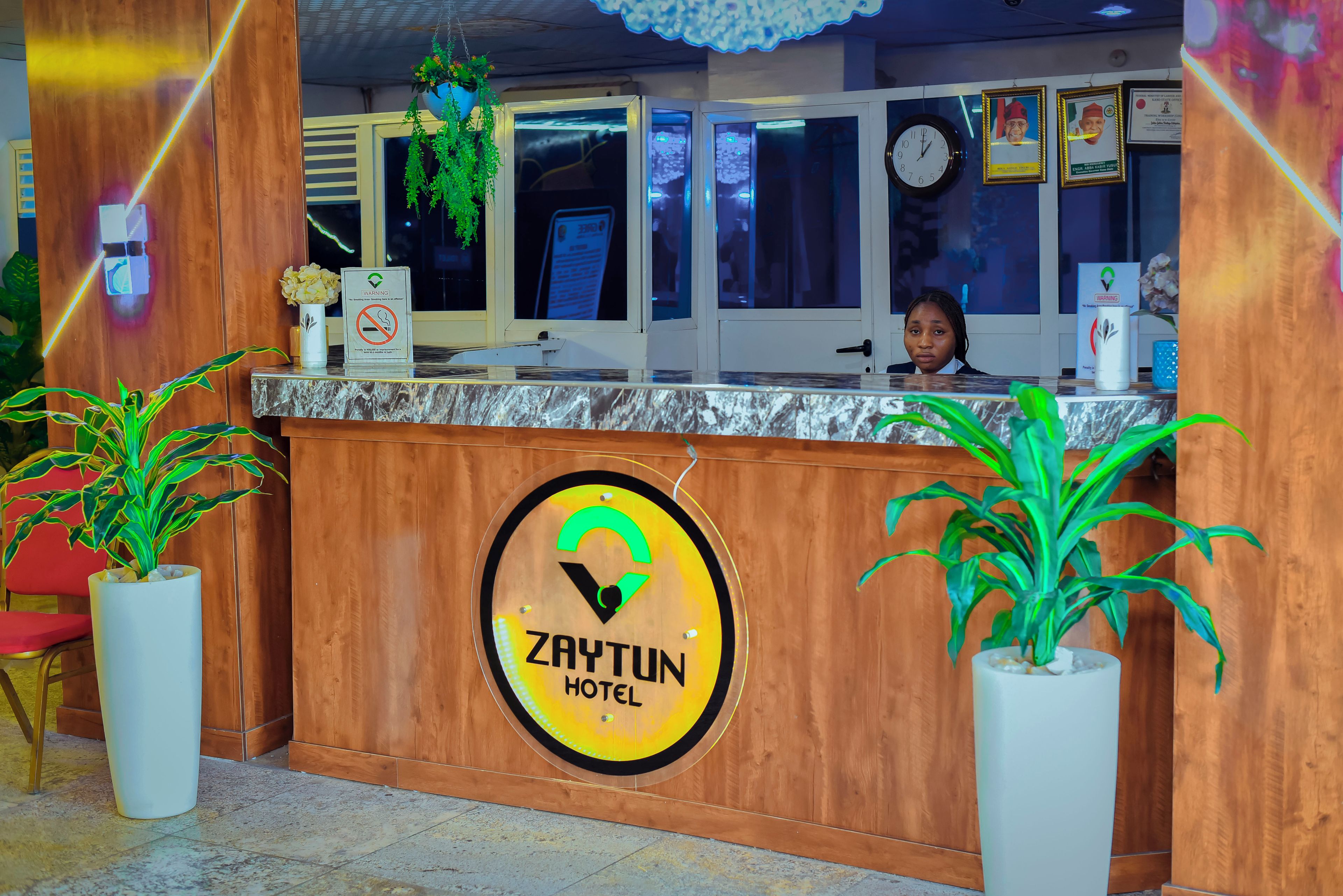 Una mesa de recepción con un letrero del Hotel Zaytun, un reloj y certificados enmarcados en la pared.