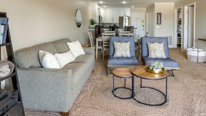 Ein Wohnzimmer mit grauem Sofa, zwei blauen Sesseln, einem Couchtisch und einer Küche im Hintergrund.
