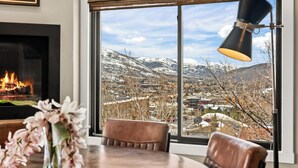 A room with a fireplace, a dining table, and a large window offering a view of a snowy landscape. - Walk to Main Street, Ontario Adventures, Two Homes Combined  - 6 BR, 2 Private Decks with Hot Tubs (Park City)