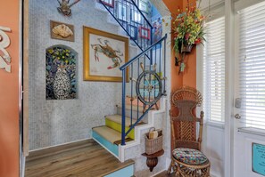 A staircase with a blue railing, a wicker chair with a colorful cushion, and a wall decorated with framed artwork and a ship's wheel.