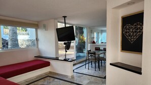 Une pièce moderne avec un banc rouge, un téléviseur à écran plat, un coin repas avec une table et des chaises, et une œuvre d’art géométrique accrochée au mur.