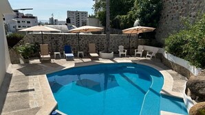 Piscina con sillones de descanso y sombrillas, rodeada de muros de piedra y follaje.