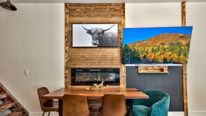 A modern dining area with a wooden table, two chairs, a fireplace, and a TV mounted on a wooden wall.