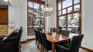 Een eetgedeelte met een houten tafel en stoelen, grote ramen met uitzicht op besneeuwde bomen, en een kroonluchter.