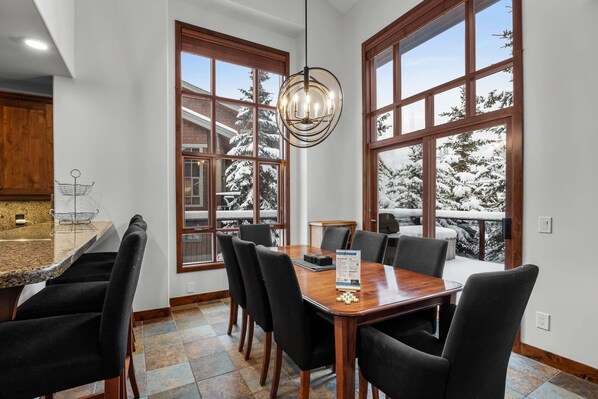 A dining area with a wooden table and chairs, large windows with a view of snow-covered trees, and a chandelier.