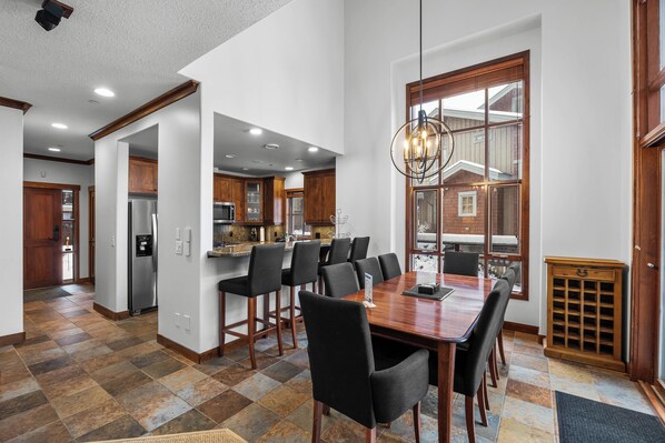 A spacious dining area with a large wooden table and black chairs, a kitchen with wooden cabinets, and a large window providing natural light.