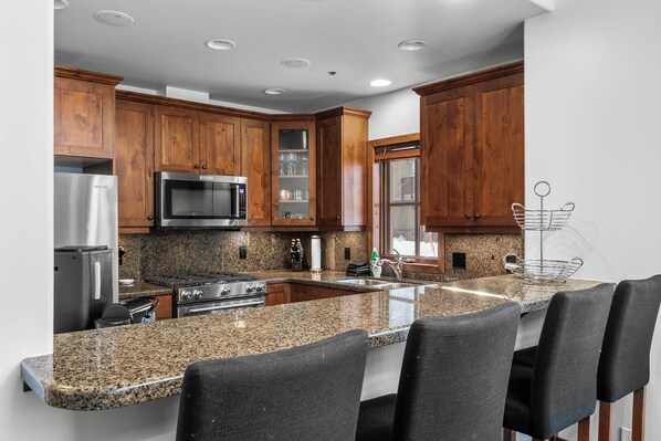 Una cocina moderna con encimeras de granito, armarios de madera, electrodomésticos de acero inoxidable y una barra desayunadora con cuatro sillas.
