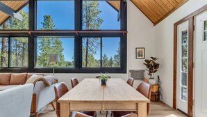 Ein geräumiges Zimmer mit einem großen Esstisch aus Holz, einer Couch und Blick auf Bäume durch große Fenster.