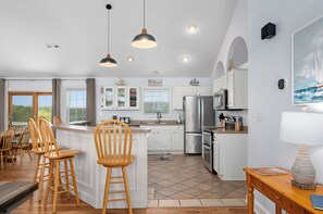 A modern kitchen with white cabinets, a central island, bar stools, pendant lighting, and a large window. - Heaven Sent: Oceanside, private pool, hot tub, rec room and EASY beach access! (Corolla)