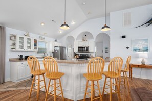 A spacious kitchen with white cabinetry, a central island with bar stools, and a dining area with a wooden table and chairs. - Heaven Sent: Oceanside, private pool, hot tub, rec room and EASY beach access! (Corolla)